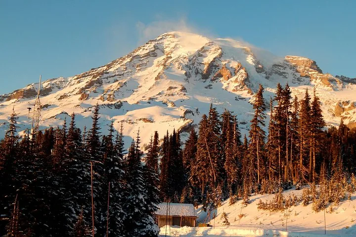 La luz del sol ilumina el Monte Rainier cubierto de nieve detrás de altos árboles de hoja perenne y un pequeño edificio.