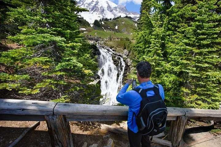 Ein Mann fotografiert einen rauschenden Wasserfall mit Mount Rainier im Hintergrund.