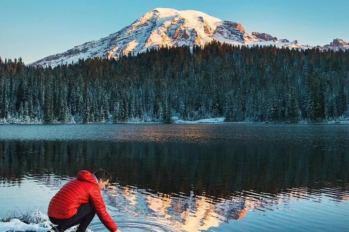 Persona agachada junto a un lago que refleja el Monte Rainier y una orilla boscosa.